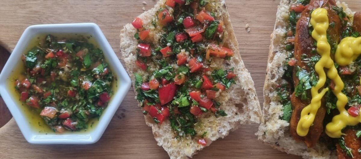 Chimichurri Vegano Receta: Salsa de chimichurri sobre una pieza de pan y sobre un choripan vegano con mostaza.