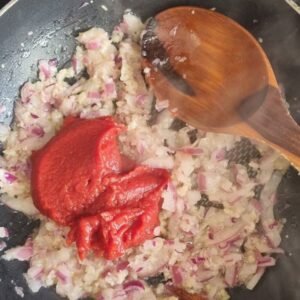 Chorizos veganos preparación paso 2: Agregar al refrito de cebollas la pasta de tomate. Cucharón de madera sobre el refrito de cebollas con pasta de tomate encima.