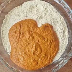 Chorizos veganos preparación paso 3: Licuar todos los ingredientes excepto el gluten hasta obtener una masa homogénea y de color intenso tomate. Liquado de los ingredienes color anaranjado sobre glutén de trigo dentro de un bowl de vidrio claro.