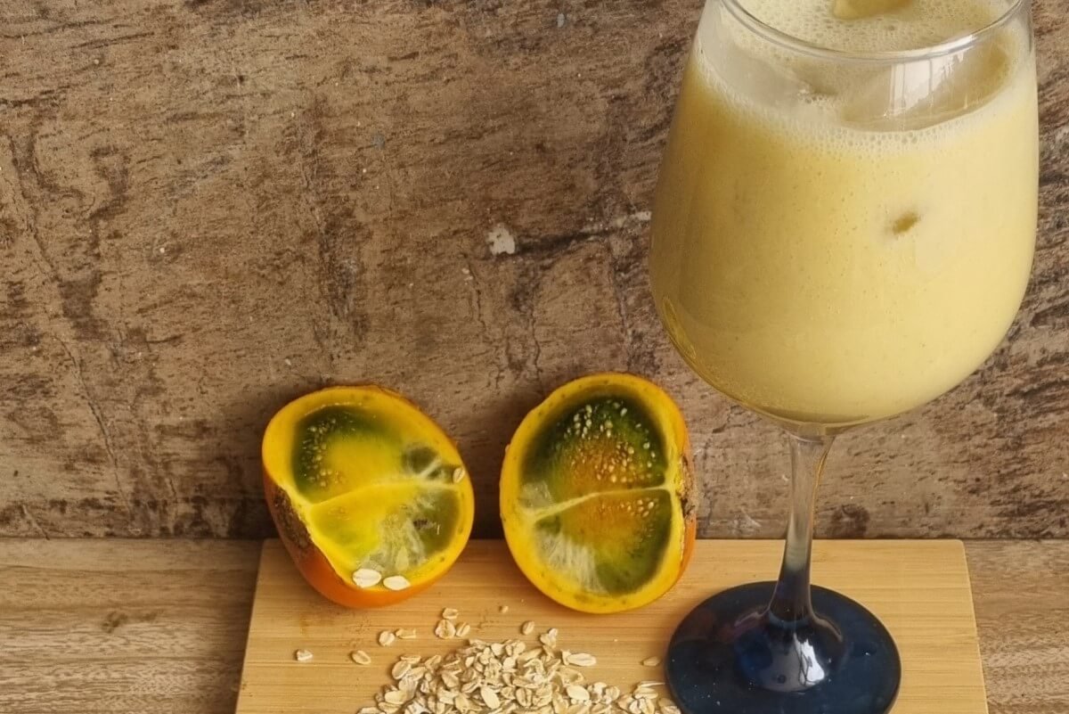 Receta de colada de avena o Quaker de naranjilla. Una copa con nuestra colada de avena, una naranjilla partida al lado izquierdo y unos granos de avena en la parte inferior.