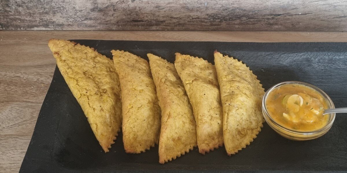 Empanadas veganas de plátano verde en una bandeja, al lado derecho un bowl con un aderezo de acompañamiento.