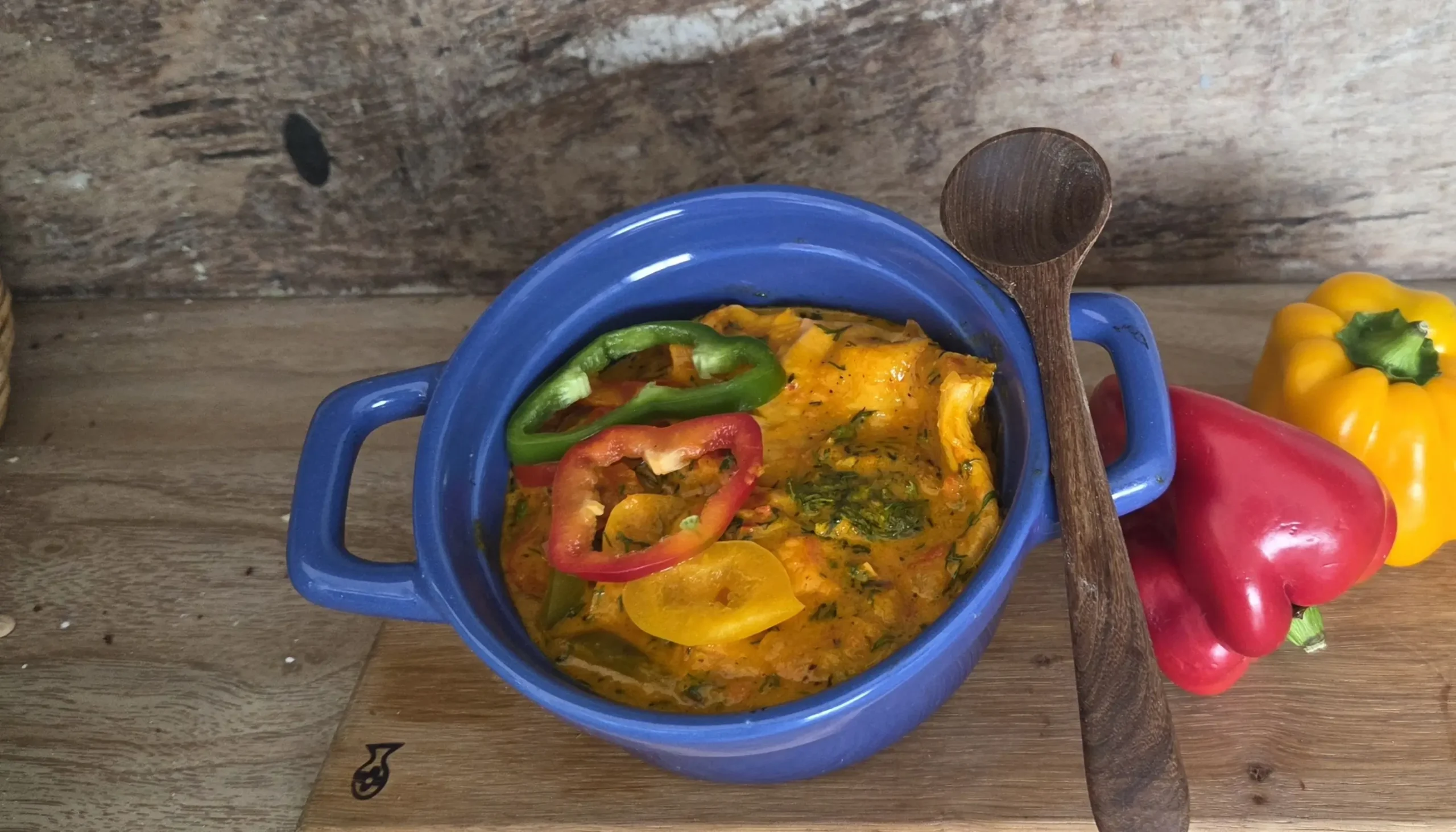Plato final de moqueca vegana servido en una cazuela azul con asas, decorado con rodajas de pimientos y cilantro picado, acompañado de una cuchara de madera al lado.